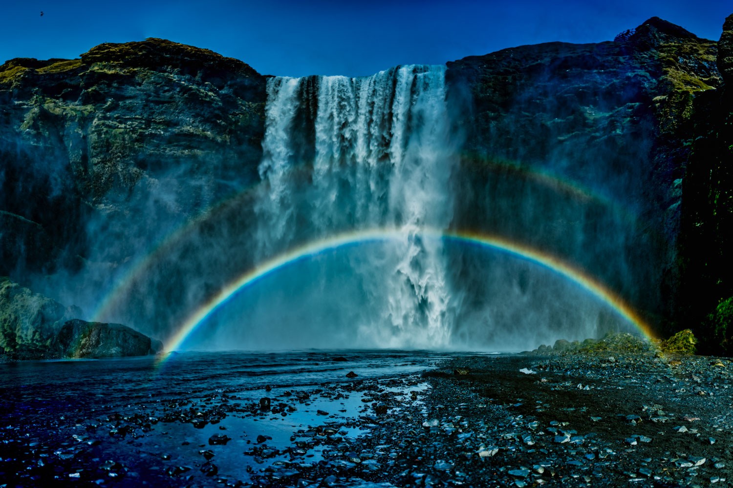 Towering Skogafoss waterfall with double rainbow and a misty rock formation. A blue sky reflected on the Skoga River with various...