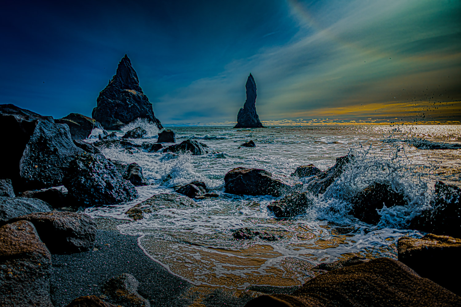 Colorful sunset with a brilliant ocean splash from the North Atlantic Ocean. Various rocks on the black sand beach including...